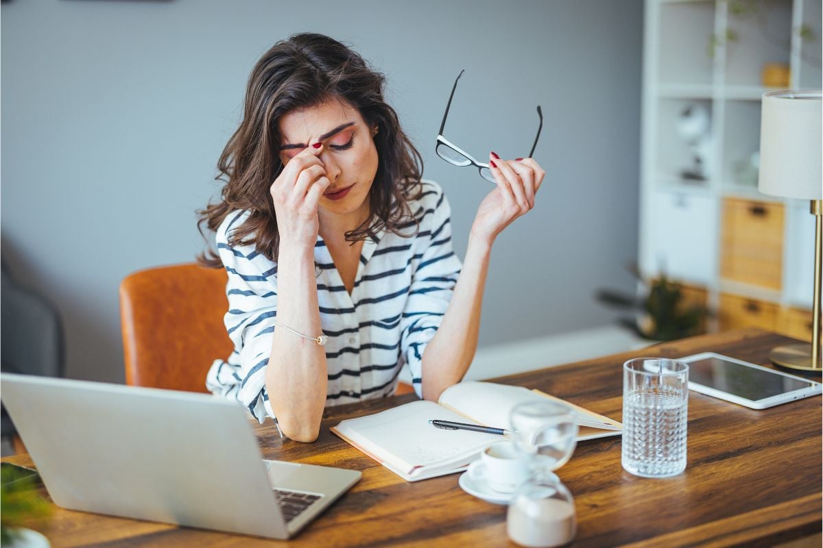 Vrouw aan tafel met laptop en papieren die bril afzet en hoofd vastpakt