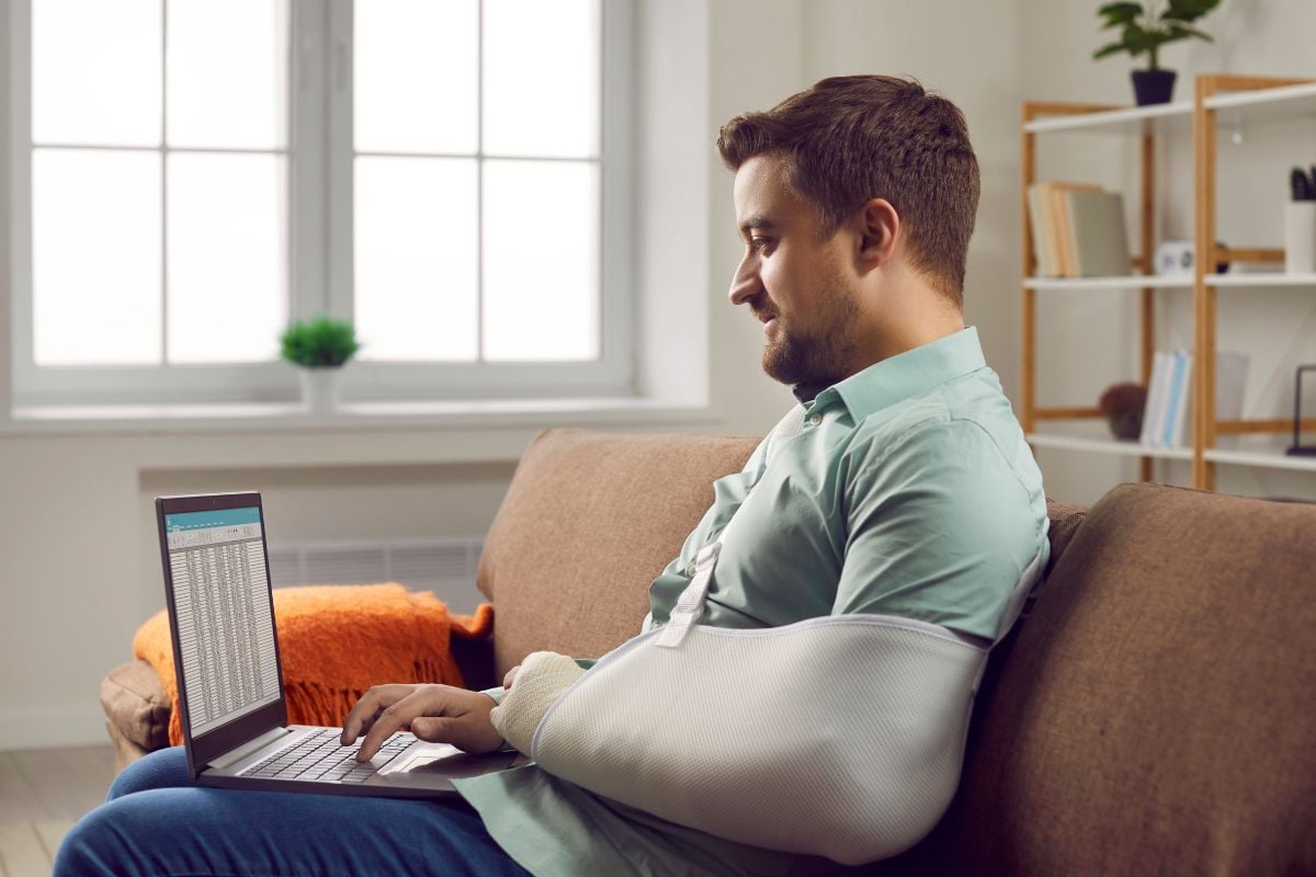 Man die op de bank zit met laptop op schoot en arm in het gips