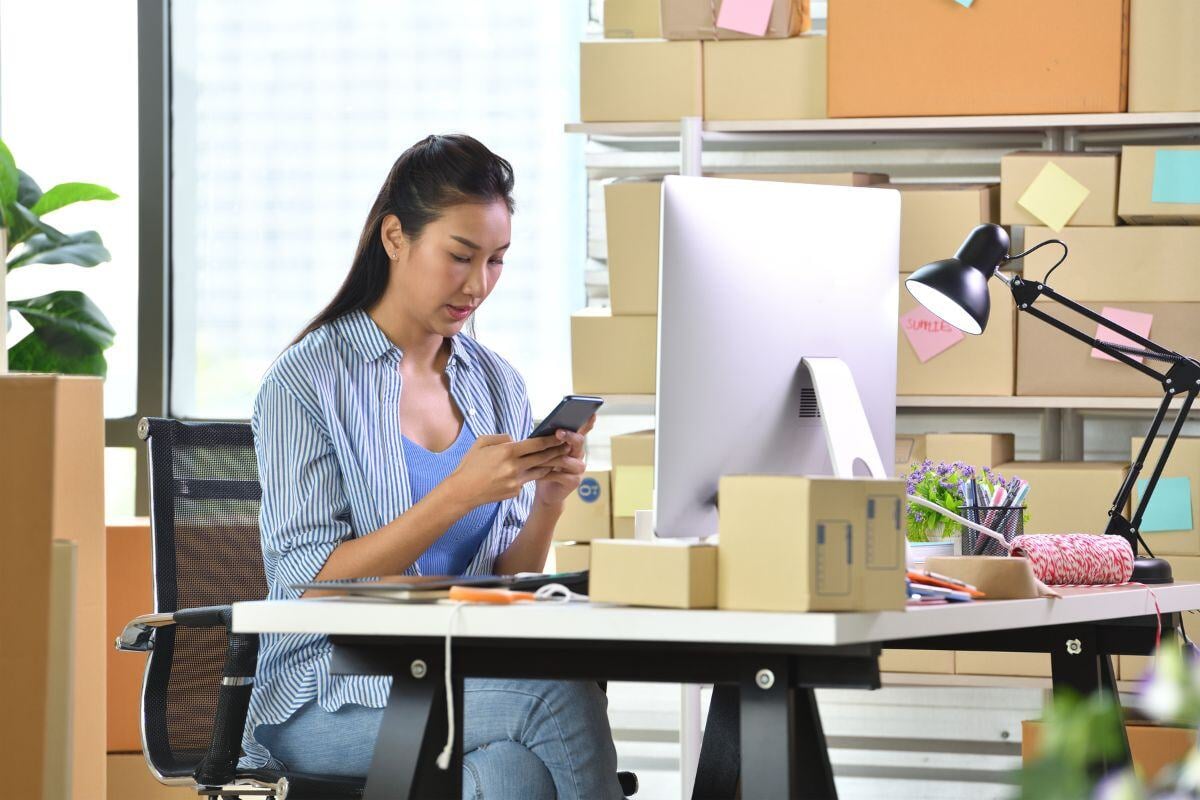 Vrouw achter computer die op haar telefoon kijkt.  Ze zit op een kantoor met allemaal pakjes om zich heen.