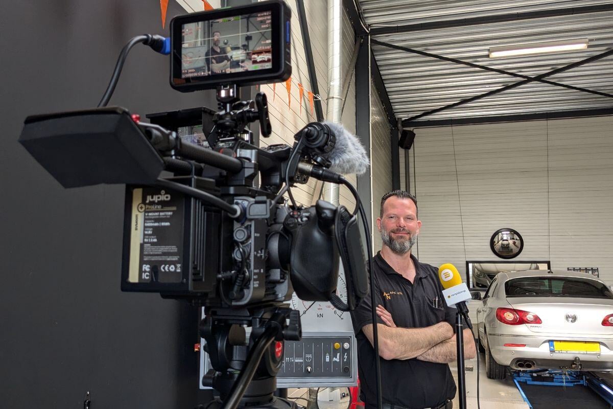 Een man staat voor een camera in een autogarage, klaar voor een interview. Hij draagt een zwart shirt en kijkt lachend in de camera. Op de achtergrond is een auto te zien die op een hefbrug staat.