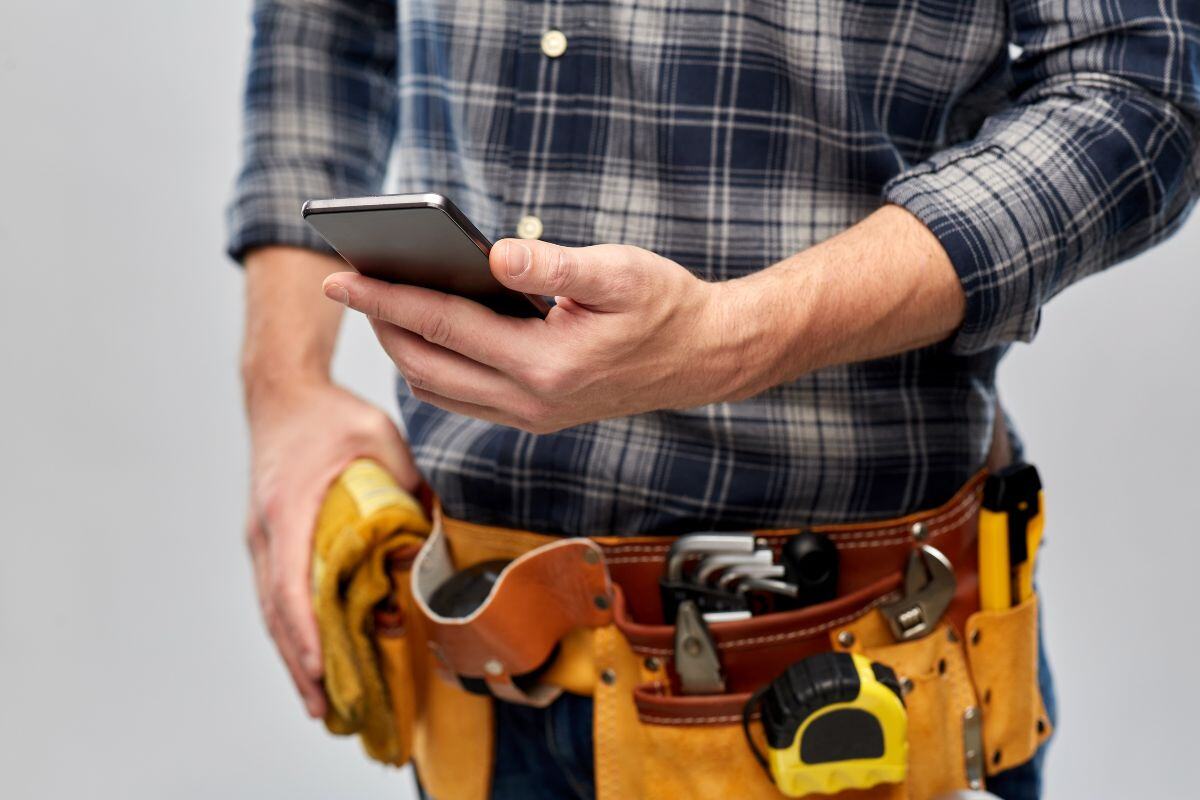 Een man in een geruite blouse gebruikt een smartphone terwijl hij gereedschap in een riem draagt. Hij houdt een werkhandschoen vast en lijkt zich klaar te maken voor een klus.