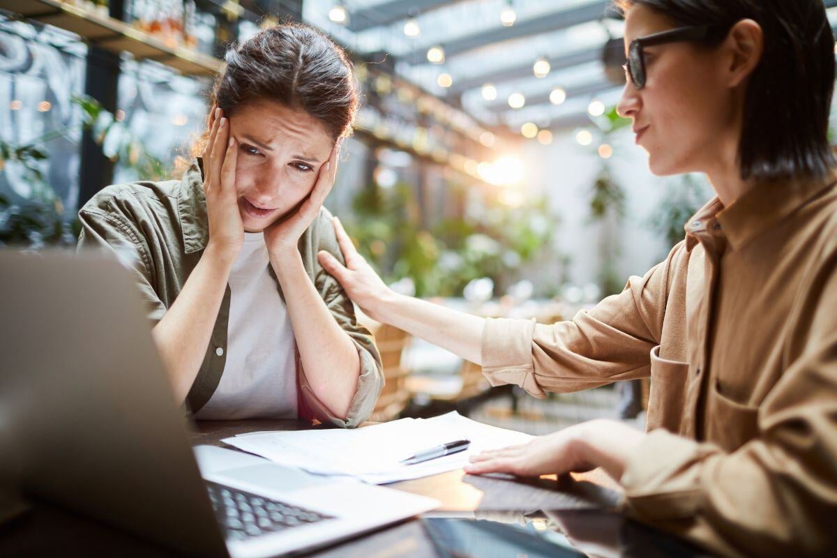 Vrouw die overstuur is aan een tafel met papieren die kijkt naar een computer. Haar adviseur steunt haar. 