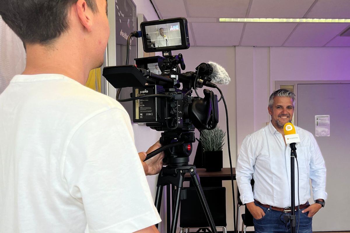 Een man in een wit overhemd staat in een kantooromgeving klaar voor de camera voor een interview. De cameraman is een klein gedeelte in beeld en er staat een gele microfoon voor de man.   