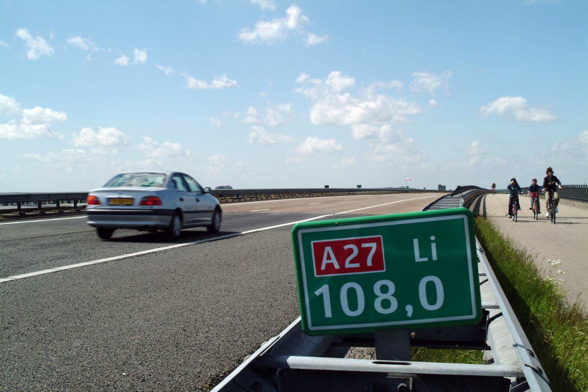 Een auto rijdt over de snelweg A27 langs kilometerpaal 108,0 terwijl drie fietsers op het naastgelegen fietspad fietsen. De lucht is helder met enkele wolken en het is een zonnige dag.