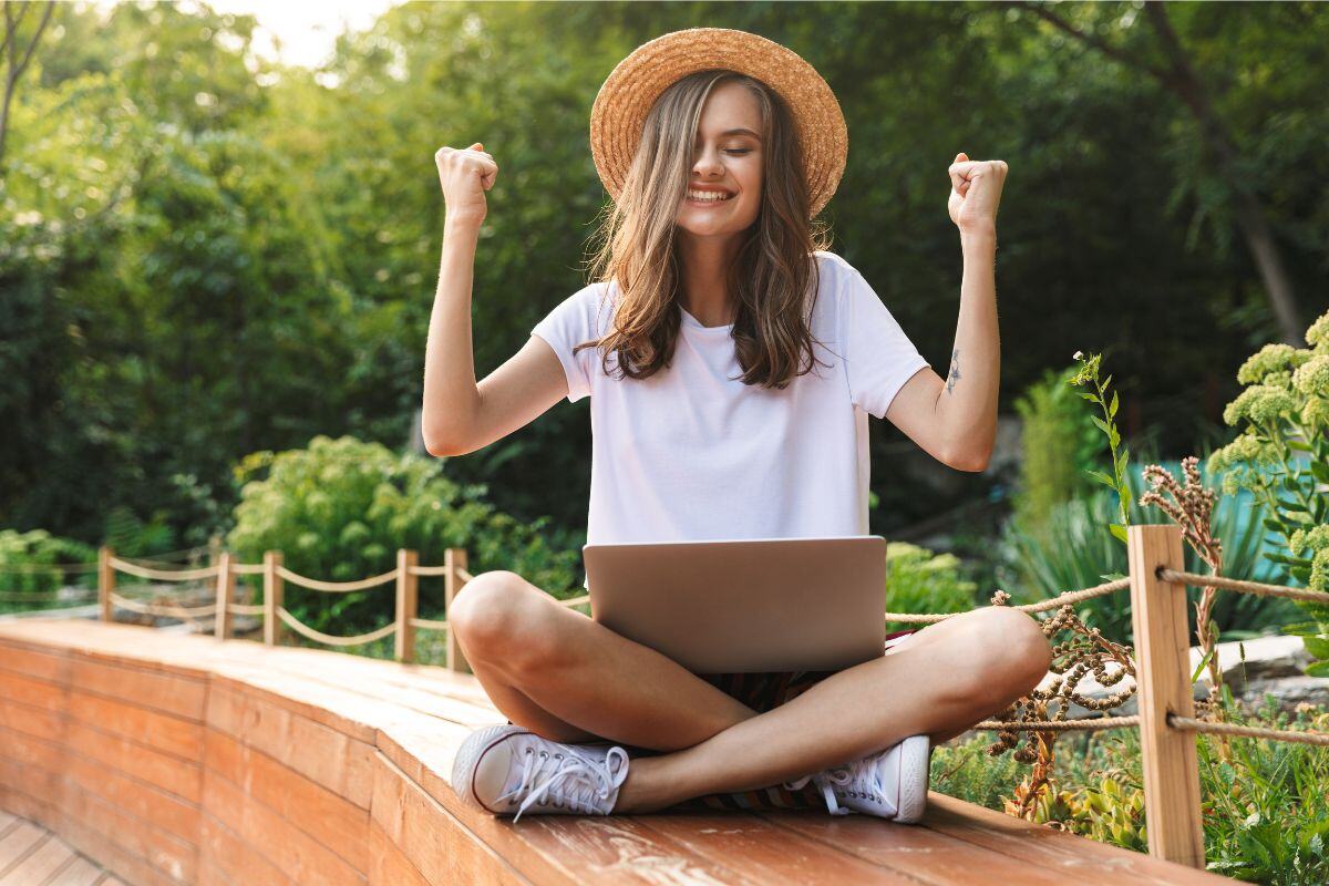 Meisje met hoed op en laptop op haar schoot die blij haar armen in de lucht steekt