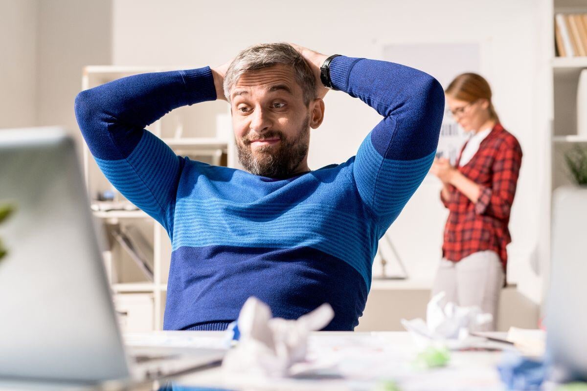 Man achter laptop met proppen papier op bureau. Met zijn handen achter zijn hoofd geslagen kijkt hij bedenkelijk naar zijn laptopscherm.